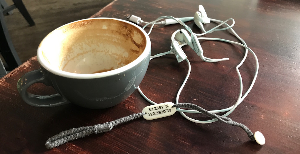 photo of an empty cappuccino cup, tangled headphones, and a woven bracelet with gps coordinates on the charm