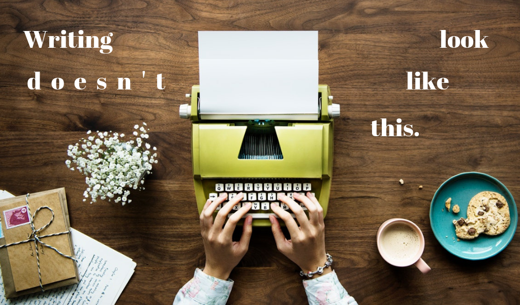 Overhead photo of a woman's hands typing on a green typewriter. To the left is an open notebook, a stack of letters, and flowers. To the right is a latte and a cookie on a blue plate. The text added to the photo reads "Writing doesn't look like this."